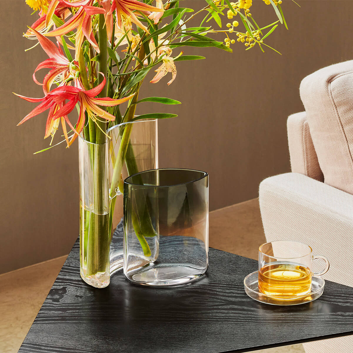 Clear glass vases with colorful flowers on a dark wooden table, with a beige sofa in the background.