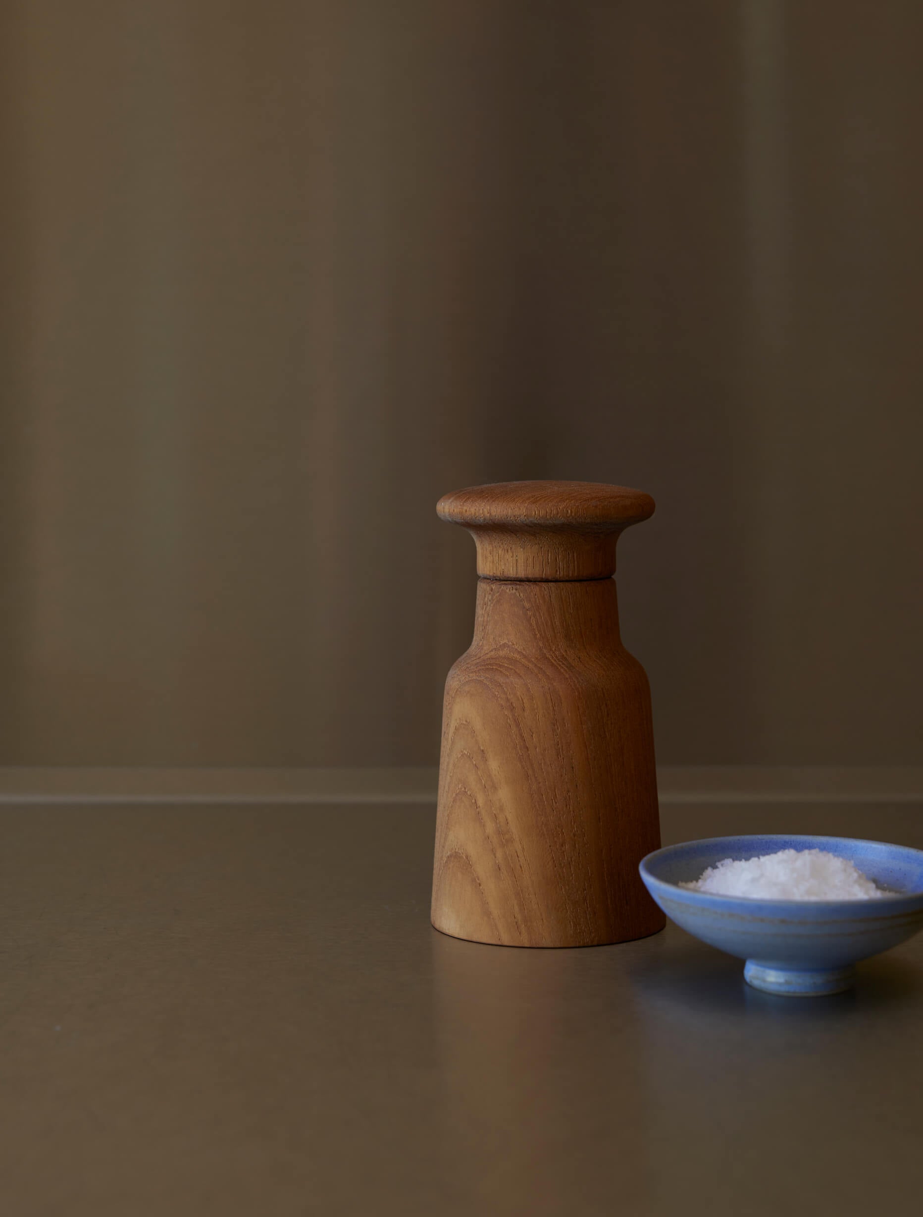 Wooden salt grinder and a small blue bowl with salt on a brown surface.