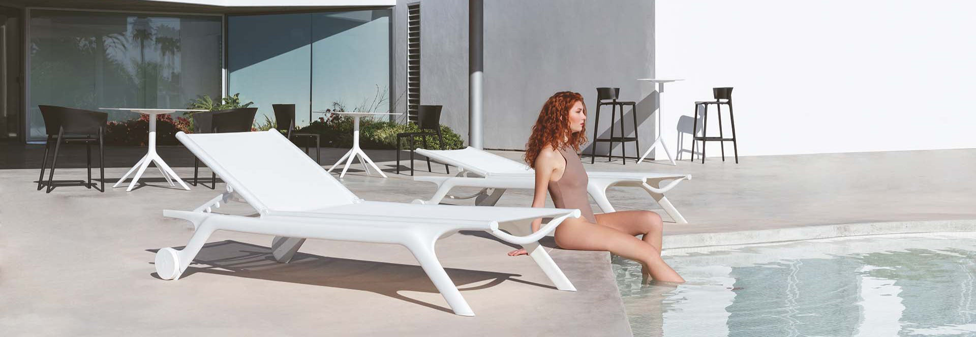 Woman sitting on a white lounge chair by a pool with modern architectural background