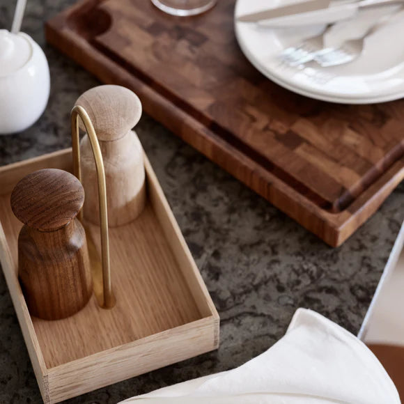 Wooden salt and pepper shakers with a gold handle on a wooden tray, placed on a dark surface.