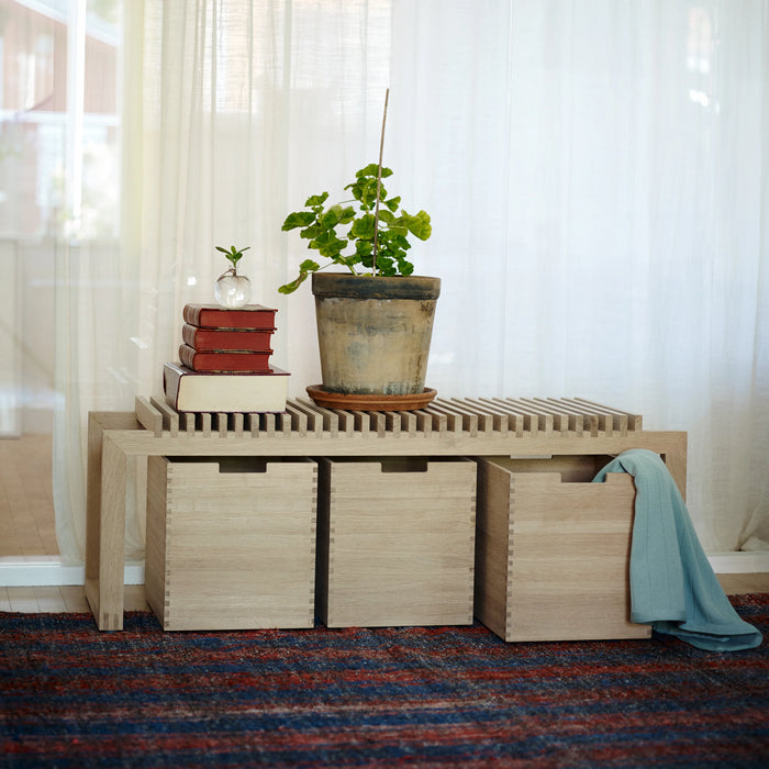 Wooden bench with storage compartments, books, and a plant in a room with white curtains.